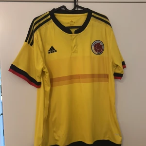 Colombia landslagströja Adidas gul - Gul Colombia fotbollströja från Adidas med svarta detaljer och ränder på axlarna. Kortärmad modell med landslagets emblem på bröstet och trycket 'UNIDOS POR UN PAÍS' på ryggen.