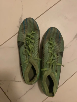 Gröna Nike Zoom fotbollsskor - Säljer ett par gröna Nike Zoom fotbollsskor med snörning och svart swoosh på sidan. Skorna har mönstrad ovandel i syntetmaterial och dobbar för bra grepp på planen. Detaljer i blått och svart ger en sportig look. Perfekta för dig som vill ta ditt spel till nästa nivå.