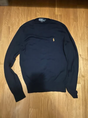 Mörkblå tröja från Polo Ralph Lauren - Klassisk mörkblå tröja från Polo Ralph Lauren med den ikoniska broderade loggan i gult på bröstet. Tröjan har rund halsringning och långa ärmar, perfekt för en clean och stilren look.