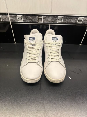 Adidas Stan Smith  - Klassiska Adidas Stan Smith sneakers i vitt skinn med marinblå detalj på hälen. Skorna har rund tå, snörning och perforerade sidor för ventilation. Ikonisk logga på plösen och yttersulan har ett mönstrat grepp. Perfekta för en clean och tidlös stil.