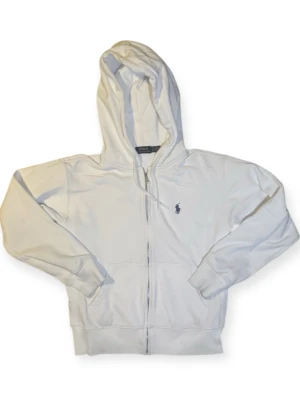 Polo Ralph Lauren vit zip hoodie - Stilren vit zip hoodie från Polo Ralph Lauren med broderad logotyp på bröstet. En klassiker som aldrig går ur stil och som funkar perfekt i en modern streetwear-garderob.  Mjuk och bekväm med huva och dragkedja framtill. Lätt att matcha med jeans, cargobyxor eller mjukisbyxor för en clean look.