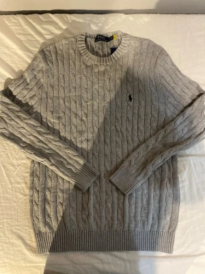 Grå kabelstickad tröja Polo Ralph Lauren - Kabelstickad grå tröja från Polo Ralph Lauren med rund halsringning och klassisk broderad logga på bröstet. Tröjan har ribbade muddar vid ärmslut och nederkant samt lång ärm. Perfekt för dig som gillar stilren och tidlös design.