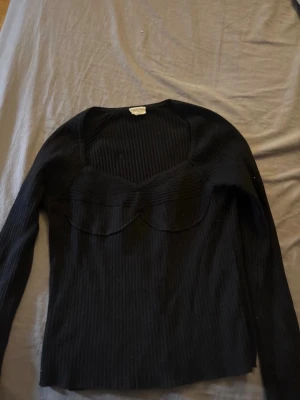 Svart ribbad långärmad topp - Säljer en svart ribbad topp med lång ärm och snygg sweetheart-urringning. Toppen har en markerad byst och är tillverkad i ett stretchigt material som sitter skönt på kroppen. Perfekt att styla med jeans eller kjol för en trendig look.