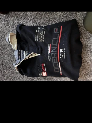 Svart pikétröja Ocean Cup 2016 svartfärgad storlek M - Säljer en svart långärmad pikétröja från Ocean Cup med broderade detaljer, amerikansk flagga och texten '2016' på bröstet. Tröjan har krage i kontrastfärg och tryck framtill. Perfekt för dig som gillar sportig stil.