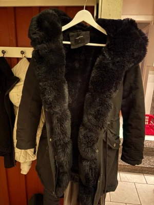 Svart parkajacka med fuskpäls - Säljer en svart parkajacka från Cypress med stor, fluffig fuskpäls längs huva och insida. Jackan har dragkedja, knappar och praktiska fickor framtill. Perfekt för kalla dagar och riktigt snygg till vintern.