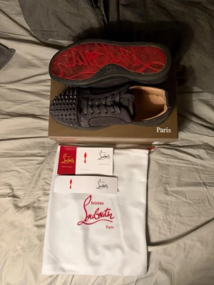 Christian Louboutin svarta sneakers med nitar - Grymma svarta sneakers från Christian Louboutin med ikonisk röd sula och coola nitar framtill. Skorna har snörning och är tillverkade i skinn med detaljer i mocka. Kommer med originalkartong, dustbag och äkthetskort. Perfekta för dig som vill sticka ut.