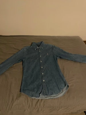 Blå jeansskjorta från Polo Ralph Lauren - Säljer en klassisk blå jeansskjorta från Polo Ralph Lauren i slim fit. Skjortan har långa ärmar, knappar framtill och den ikoniska broderade loggan på bröstet.