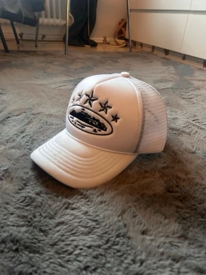 Vit meshkeps från Corteiz - Säljer en vit truckerkeps från Corteiz med svart broderad logga och stjärnor framtill och på sidan. Kepsen har meshpaneler runtom för extra ventilation och justerbar snapback baktill. Snygg och clean design med Corteiz-band på insidan.