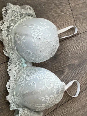Spets BH - Bilderna visar en ljusblå spets-bh med blommigt spetsmönster och liten rosett framtill, inte en väst.