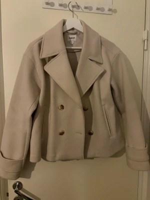 Beige kappa från Kappahl - Säljer en beige kappa från Kappahl med dubbelknäppning. Perfekt för dig som vill ha en stilren och tidlös look.