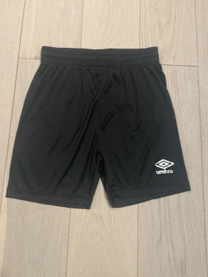 Svarta träningsshorts från Umbro - Säljer ett par klassiska svarta träningsshorts från Umbro. De har elastisk midja och är tillverkade i ett lätt och svalt syntetmaterial. Perfekta för fotboll eller annan träning. Umbro-logga i vitt på både fram- och baksidan av shortsen.