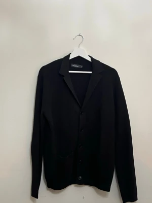 Svart cardigan jacka från Dressmann - Säljer en svart cardigan jacka från Dressmann med klassisk krage och knäppning framtill. Jackan har två fickor fram och är långärmad. Perfekt för dig som gillar stilrena och enkla plagg.