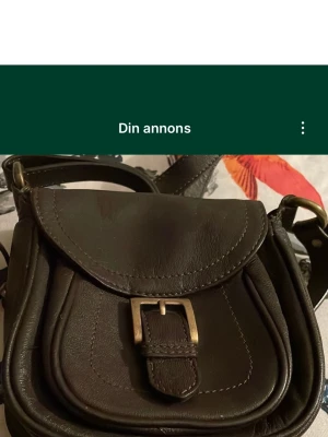 Brun axelväska i skinn med spänne - Snygg brun axelväska i skinn med klassiskt spänne framtill. Väskan har rundad form och justerbar axelrem. Perfekt storlek för det viktigaste och stilren design som passar till många outfits. Två fack inne i väskan varav en med dragkedja. Stängs med magnetknapp. Säljs på fler sidor.