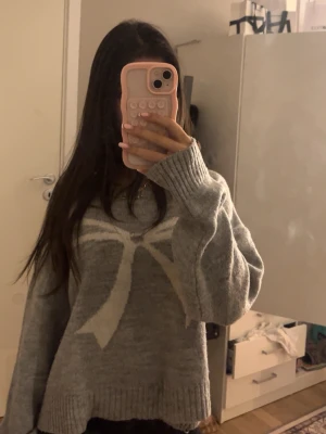 Grå stickad tröja med rosettmönster - Mysig grå stickad tröja med oversized passform och lång ärm. Tröjan har ett stort vitt rosettmönster framtill och breda ribbade muddar vid ärmslut och nederkant. Perfekt för kalla dagar och enkel att styla till jeans eller kjol.