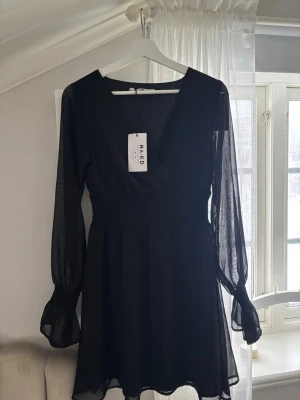 Svart omlottklänning från NA-KD - Säljer en svart kort omlottklänning från NA-KD med v-ringning och långa, transparenta ärmar. Klänningen har smock i midjan och vid ärmsluten för en snygg passform. Perfekt för dig som gillar stilrena och trendiga plagg.