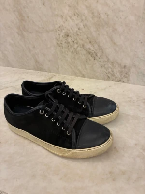 Lanvin  - Säljer ett par klassiska mörkblå sneakers från Lanvin med vit sula och svarta snören. Skorna har rund tå, platt sula och detaljer i mocka och skinn. Perfekta till jeans eller chinos för en clean look.