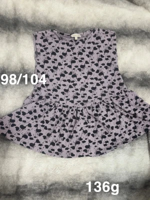 Lila blommig tunika från pompdelux  - Supersöt lila tunika från Pompdelux med svart blommigt mönster. Tunikan har ärmlös design och en volang nedtill som ger en extra söt touch. Perfekt för dig som gillar lekfulla och unika plagg!🌸Fint använt skick. Storlek 98/104