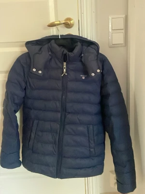 Mörkblå pufferjacka från Gant - Säljer en mörkblå pufferjacka från Gant med huva och dragkedja framtill. Jackan har quiltad design, praktiska fickor och snygga detaljer med knappar vid huvan. Perfekt för kalla dagar och riktigt skön att bära.