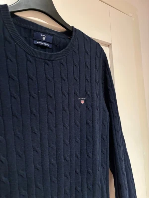 Marinblå kabelstickad tröja från GANT - Kabelstickad marinblå tröja från GANT i stretchig bomull. Tröjan är i helt nytt skick, jätte mjuk och mysig, samtidigt som den strimlar elegans.
