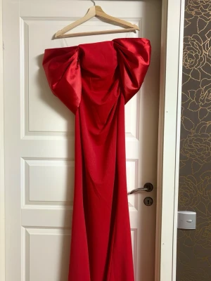 Röd axelbandslös maxiklänning med rosett - Säljer en statement röd långklänning med stor rosett baktill. Klänningen är axelbandslös och har en elegant, svepande siluett. Perfekt för dig som vill sticka ut på bal eller fest!