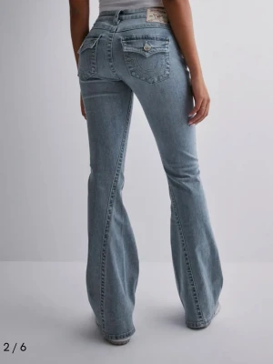 True Religion Joey Low Rise Flare Jeans - Snygga ljusblå jeans från True Religion, modell Joey Low Rise Flare. De har låg midja, utsvängda ben och klassiska fickor med lock och silverknappar. Jeansen är gjorda i mjukt denim och har tydliga sömmar och ikonisk logga bak.