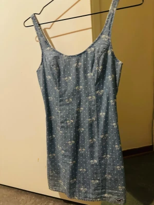 Blå kortklänning från Hollister - Supersöt blå kortklänning från Hollister med vita palmmönster och prickar. Klänningen har breda axelband, rundad urringning och dragkedja i ryggen. Tillverkad i mjuk bomull, perfekt för sommarens alla dagar.