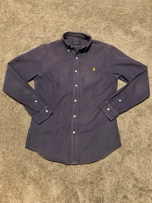Mörkblå skjorta från Polo Ralph Lauren - Klassisk mörkblå skjorta från Polo Ralph Lauren i slim fit. Skjortan har långa ärmar, knappar framtill och gul broderad logga på bröstet. Tillverkad i mjuk bomull och passar perfekt till en clean och stilren look.