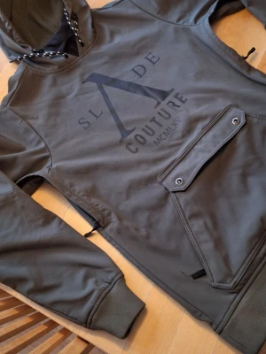 Grön skidjacka från Slade Couture XS - Säljer en grön skidjacka från Slade Couture i storlek XS. Jackan har huva med snörning, stor ficka framtill med tryckknappar och ribbade muddar. Stora logotryck både fram och bak.