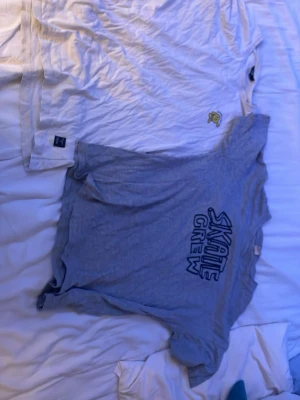Två skateinspirerade t-shirts - Säljer två t-shirts med skatevibe. Ena är vit med tryck av en gul figur på bröstet och texten 'STAY TRUE STAY YOU' på ryggen. Den andra är ljusgrå med 'SKATE CREW' tryck fram och ett stort grafiskt tryck bak. Båda har rund hals och korta ärmar, perfekta för chill dagar.