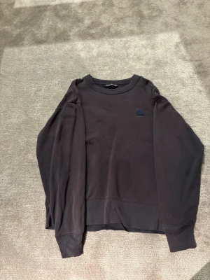 Acne Studios Tröja - En stilren marin blå sweatshirt från Acne Studios med rund halsringning och diskret logopatch på bröstet. Tillverkad i mjuk bomull och har långa ärmar samt ribbade muddar vid ärmslut och nederkant. Perfekt för en clean och avslappnad look. (Finns två små hål)