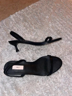 Svarta sandaler med klack från Nelly - Säljer ett par stilrena svarta sandaler från Nelly med smal rem över tårna och runt hälen. Skorna har låg klack och öppen tå, perfekta till sommarens outfits. 