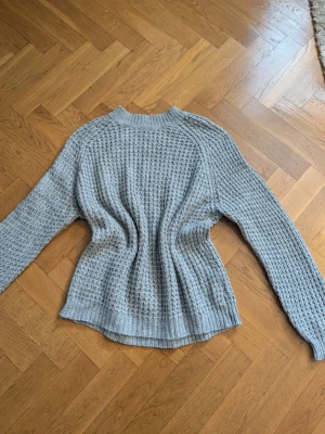 Grå stickad tröja från 157 - Mysig grå stickad tröja från 157 i storlek M. Tröjan har ett grovt, strukturerat stickmönster och ribbade muddar vid hals, ärmslut och nederkant. Perfekt för kyliga dagar och enkel att matcha med jeans eller kjol.