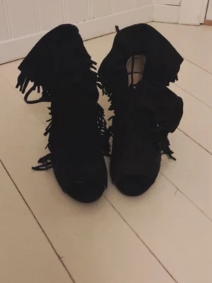 Svarta mocka boots med fransar och klack - Säljer ett par svarta boots i mocka med hög klack och öppen tå. Skorna har snygga fransdetaljer längs sidorna och snörning framtill. Perfekta för dig som vill sticka ut med en cool och trendig look.
