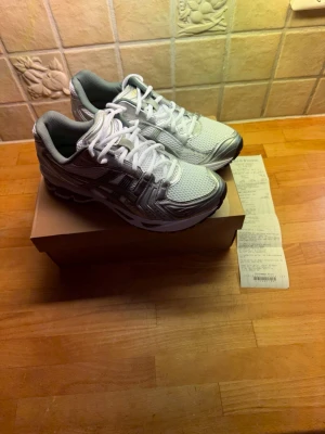 Asics Gel-Kayano 14 sneakers vit/silver - Säljer ett par Asics Gel-Kayano 14 sneakers i vitt och silver med gröna detaljer. Skorna har mesh- och syntetmaterial, klassisk snörning och en chunky sula för maxad komfort. Perfekta för dig som gillar retrovibbar och sportig stil.