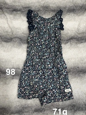 Blommig byxdress från Newbie - Supersöt blommig byxdress från Newbie i mörkblått med små blommor i pastellfärger. Byxdressen har volangdetaljer vid axlarna. Perfekt för varma dagar och enkel att ta på.🌸 Fint använt skick. Storlek 98. 