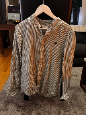 Randig henley-skjorta från Pier One - Säljer en randig henley-skjorta från Pier One i bomull. Skjortan har smala svarta och vita ränder, knappar framtill och lång ärm. Klassisk ståkrage och liten broderad logga på bröstet. Perfekt för dig som gillar en chill och stilren look.