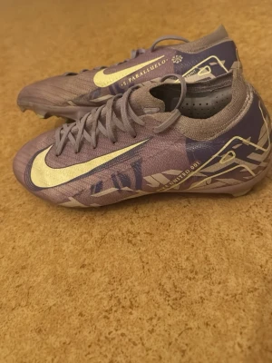 Nike fotbollsskor lila/guld - Säljer ett par Nike fotbollsskor i lila med guldiga detaljer och snörning. Skorna har en snygg, sportig design med mönstrade inslag och stor Nike-logga på sidan. Ovandelen är i syntetmaterial och skon har en smal passform som ger bra bollkontroll.