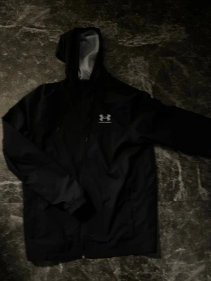 Under armour vindjacka  - Säljer en svart vindjacka från Under Armour med huva och dragkedja framtill. Jackan har en enkel, sportig design med logga på bröstet och justerbar huva med snörning. Perfekt för blåsiga dagar och outdoor-aktiviteter.