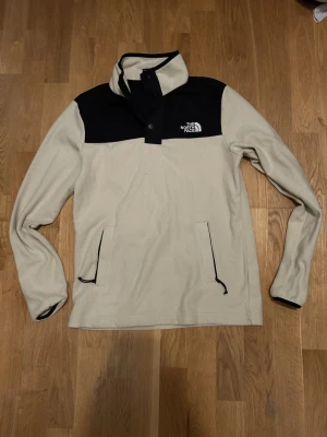 Beige fleece från The North Face - Säljer en beige och svart fleece från The North Face med hög krage och knappstängning. Tröjan har två fickor med dragkedja och logga på bröstet. Perfekt för kyliga dagar och riktigt skön att ha på sig.