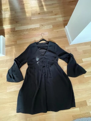 Svart klänning med v-ringning - En svart klänning med v-ringad hals och vida, långa ärmar. Klänningen har en lätt transparent look och är sydd i ett tunt, flowy material