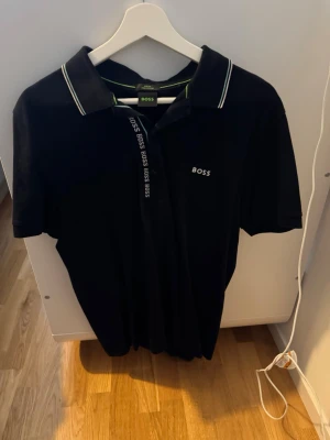 Svart piké från BOSS med logga - Säljer en svart pikétröja från BOSS i slim fit med klassisk krage och vita samt gröna detaljer på kragen. Pikén har BOSS-logga broderad på bröstet och knäppning framtill med BOSS-tryck. Tillverkad i mjuk pima bomull för extra komfort.