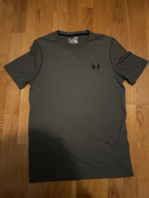Mörkgrå t-shirt från Under Armour - Säljer en mörkgrå t-shirt från Under Armour med klassisk logga på bröstet. T-shirten har korta ärmar och är gjord i ett mjukt och lätt material som passar perfekt till träning. Enkel och stilren design som funkar till det mesta.