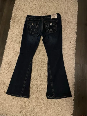 Mörkblå True Religion bootcut jeans - Snygga mörkblå jeans från True Religion med klassiska kontrastsömmar och ikoniska ficklock med silverfärgade knappar. Modellen har bootcut-ben och markerade bakfickor med broderad logga. Perfekt för dig som gillar statement-jeans med coola detaljer.