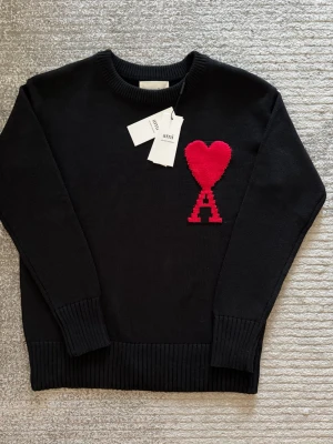 Svart stickad tröja från Ami - Svart stickad tröja från Ami Alexandre Mattiussi med ikonisk röd hjärtdetalj och bokstaven A på bröstet. Klassisk rund halsringning och ribbade muddar vid ärmar och nederkant. Perfekt för dig som vill ha en clean men ändå statement look.