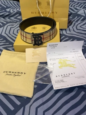 Rutig Burberry bälte i skinn - Säljer ett klassiskt Burberry bälte i äkta skinn med ikoniskt rutigt mönster i beige, rött och svart. Bältet har en svart metallspänne med Burberrys logotyp. Perfekt accessoar för att lyfta din outfit.