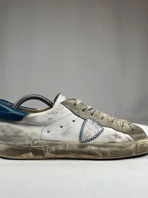 Philippe Model vita sneakers med blå detalj - Säljer ett par vita sneakers från Philippe Model med grå mockadetaljer, blå häl och broderad logga på sidan. Skorna har snörning med text på skosnörena och en platt sula. Perfekta för dig som gillar en urban och avslappnad stil.