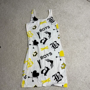 Vit kortklänning med tryck från HM Divided - Säljer en vit kortklänning från HM Divided med coolt svart och gult tryck, bland annat hjärtan, pilar och text. Klänningen är ärmlös med smala axelband och har en rak passform. Perfekt för dig som gillar statement-plagg och vill sticka ut.