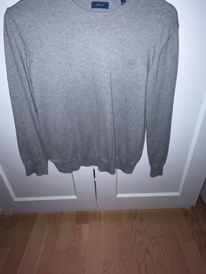 Grå stickad tröja från GANT - Säljer en stilren grå stickad tröja från GANT med rund halsringning och diskret broderad logga på bröstet. Tröjan har långa ärmar och ribbade muddar vid ärmslut och nederkant. Perfekt till lager på lager under höst och vinter.