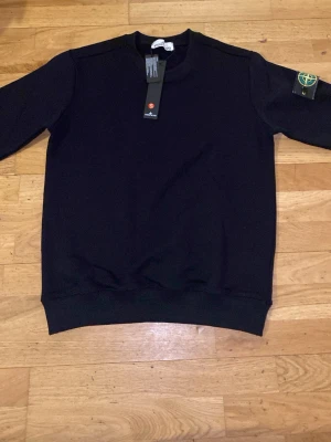 Svart sweatshirt från Stone Island - Svart sweatshirt från Stone Island med klassisk rund hals och ribbade muddar. Tröjan har den ikoniska Stone Island-loggan på vänster ärm. Tillverkad i mjuk bomull för en skön känsla och stilren look. Tröjan har aldrig använts. 