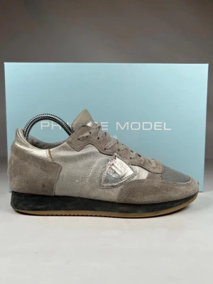 Grå sneakers från Philippe Model - Säljer ett par snygga sneakers från Philippe Model i grått och silver. Skorna har ovandel i mocka och mesh, samt detaljer i metallicskinn på sidan och tungan. Klassisk snörning och svart sula med gummidetalj. Perfekta för dig som gillar stilrena men ändå unika sneakers. Kommer med box till 